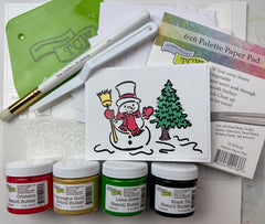 Art supplies including stencil, stencil paint jars, a brush, and a card with a snowman and tree design on a white background.